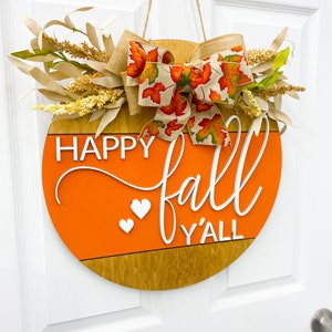 May include: A round wooden door hanger with a burlap bow and fall-themed ribbon. The hanger is painted orange and brown and has the text "Happy fall y'all" in white lettering.