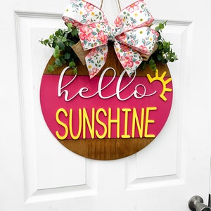 May include: A pink and brown wooden round door hanger with the text "hello sunshine" in yellow letters. A floral bow with pink and white flowers is attached to the top of the hanger.
