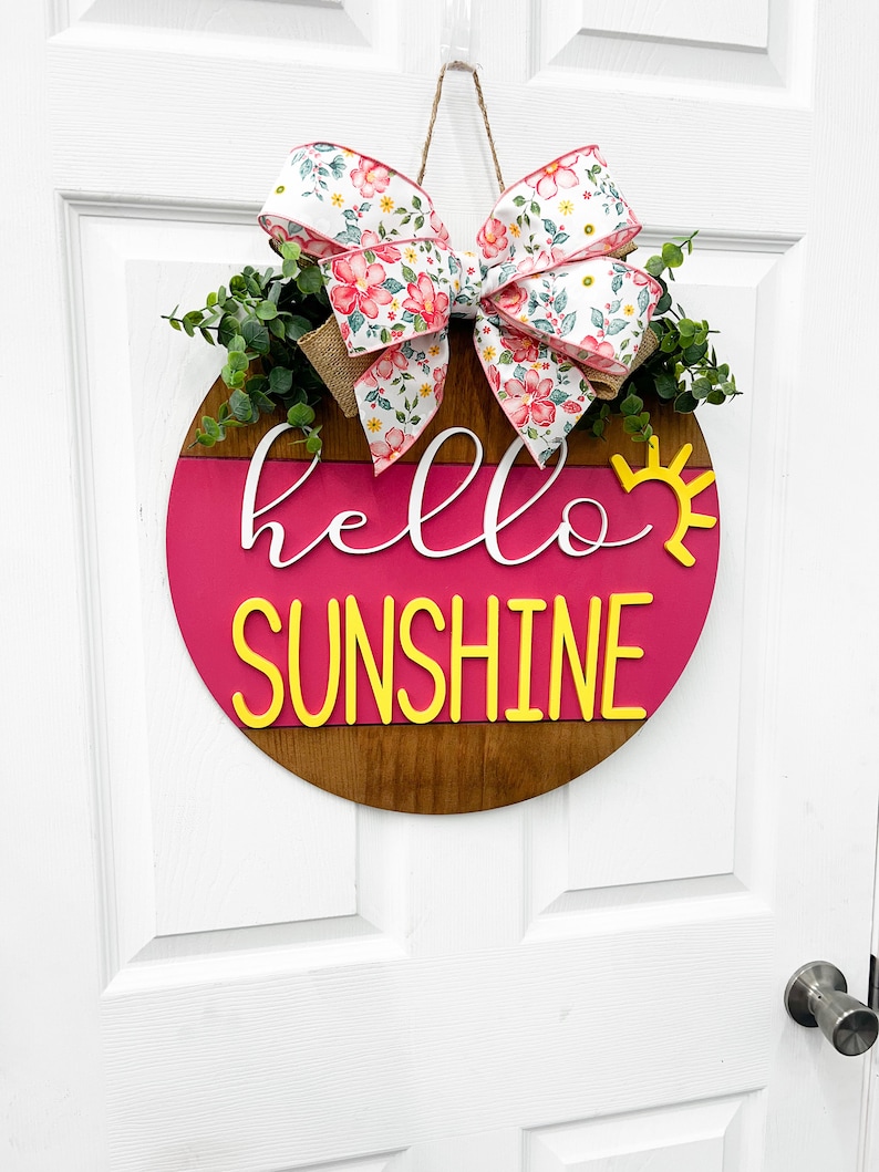 May include: A pink and brown wooden round door hanger with the text "hello sunshine" in yellow letters. A floral bow with pink and white flowers is attached to the top of the hanger.