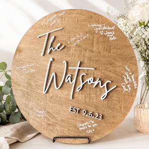 May include: A round wooden sign with the words "The Watsons" in white script. The sign has handwritten messages and the date "EST 9.6.23". The sign is displayed on a black metal stand, with flowers and a candle in the background.