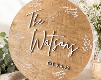 May include: A round wooden sign with the words "The Watsons" in white script. The sign has handwritten messages and the date "EST 9.6.23". The sign is displayed on a black metal stand, with flowers and a candle in the background.