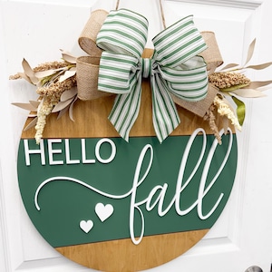 May include: A round wooden door sign featuring a dark green horizontal panel with the white 3D text HELLO fall and two small white hearts. The sign is decorated with a large bow made of green and white striped ribbon and burlap, plus beige foliage.