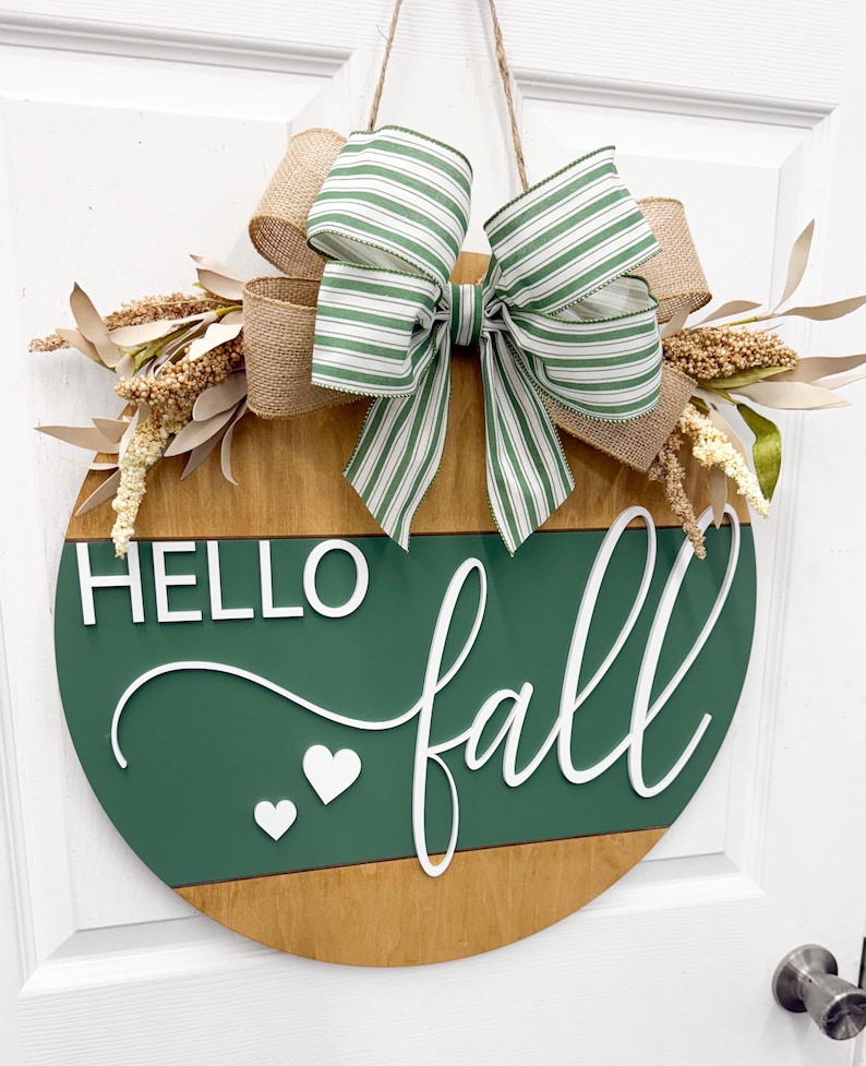 May include: A round wooden door sign featuring a dark green horizontal panel with the white 3D text HELLO fall and two small white hearts. The sign is decorated with a large bow made of green and white striped ribbon and burlap, plus beige foliage.
