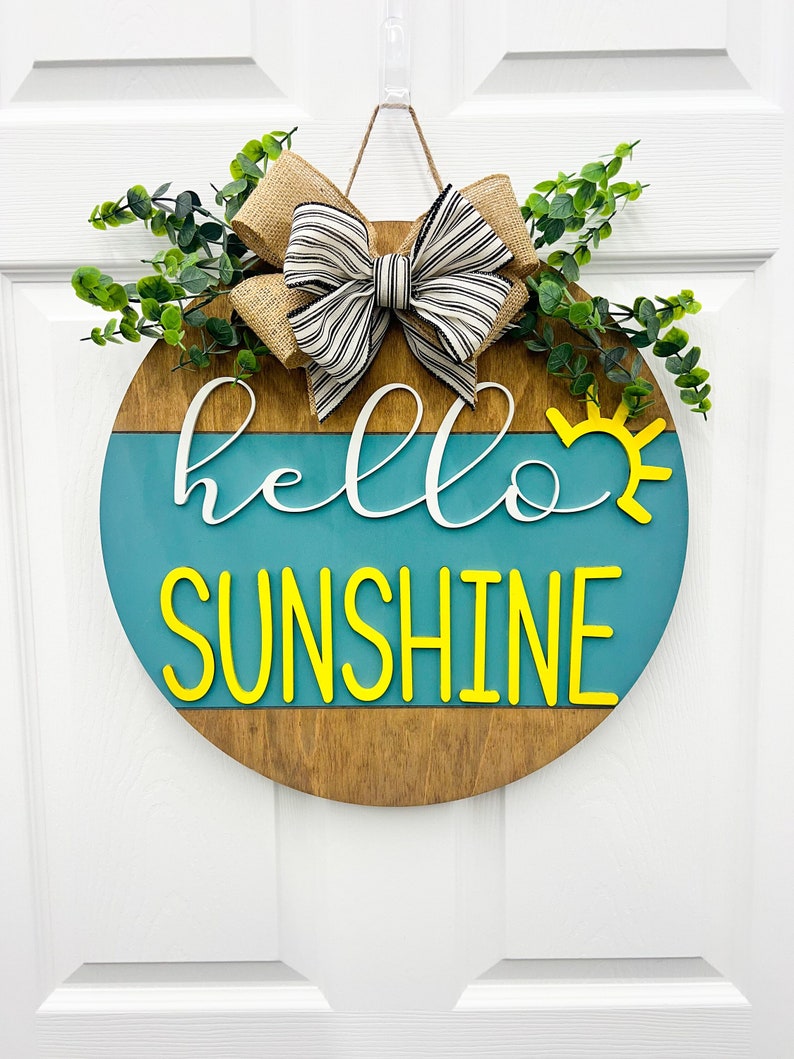 May include: A round wooden door hanger with a blue and brown background. The words "hello sunshine" are written in white and yellow letters. A burlap bow with black and white stripes is attached to the top of the hanger.
