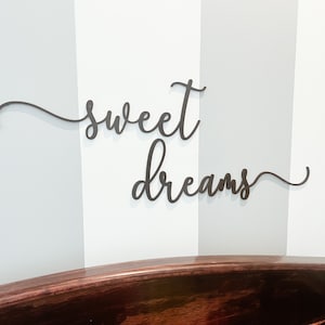 May include: A wooden sign with the words "sweet dreams" in a cursive font, mounted on a white wall with vertical stripes. The sign is above a dark brown wooden headboard.