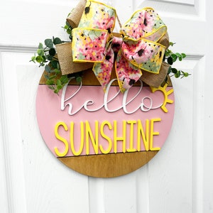 May include: A pink and brown wooden round door hanger with the text "hello sunshine" in white and yellow letters. A floral bow with pink and yellow flowers is attached to the top of the hanger.