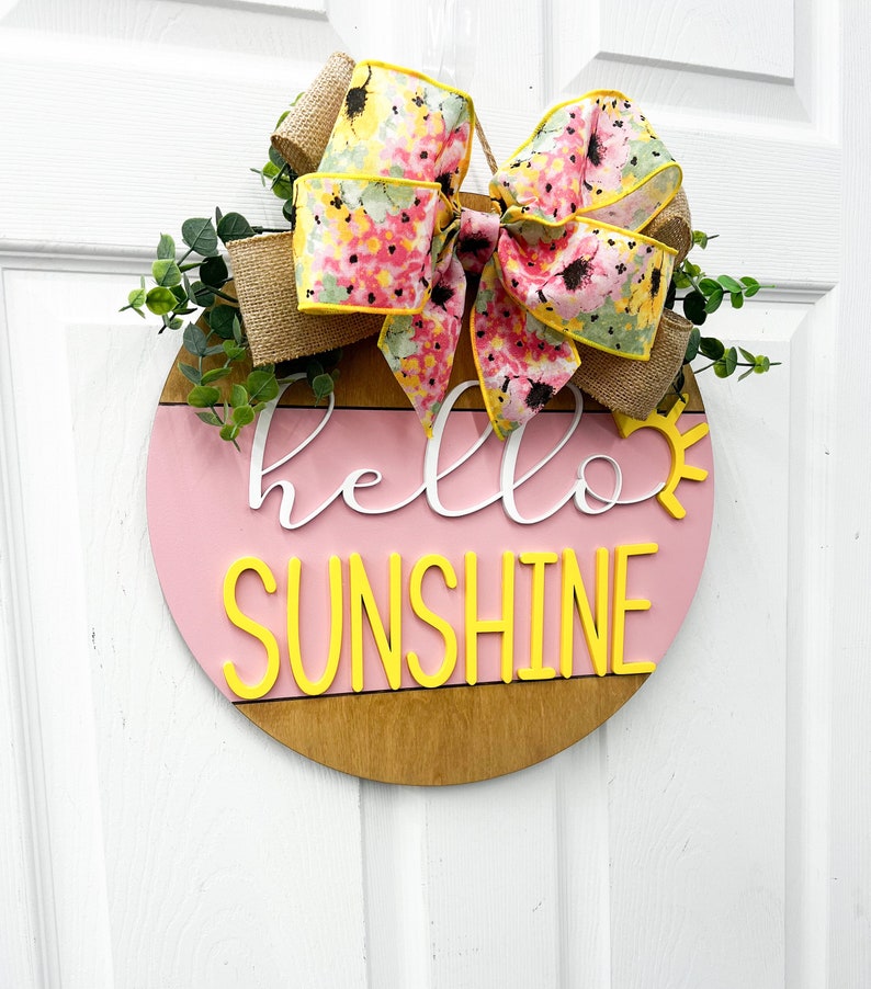 May include: A pink and brown wooden round door hanger with the text "hello sunshine" in white and yellow letters. A floral bow with pink and yellow flowers is attached to the top of the hanger.