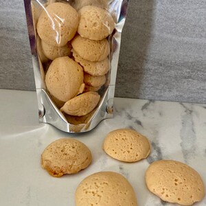 Puede incluir: Una bolsa de cierre herm&eacute;tico plateada llena de galletas redondas y esponjosas de color marr&oacute;n claro. Cuatro galletas adicionales est&aacute;n esparcidas sobre una superficie de m&aacute;rmol blanco.