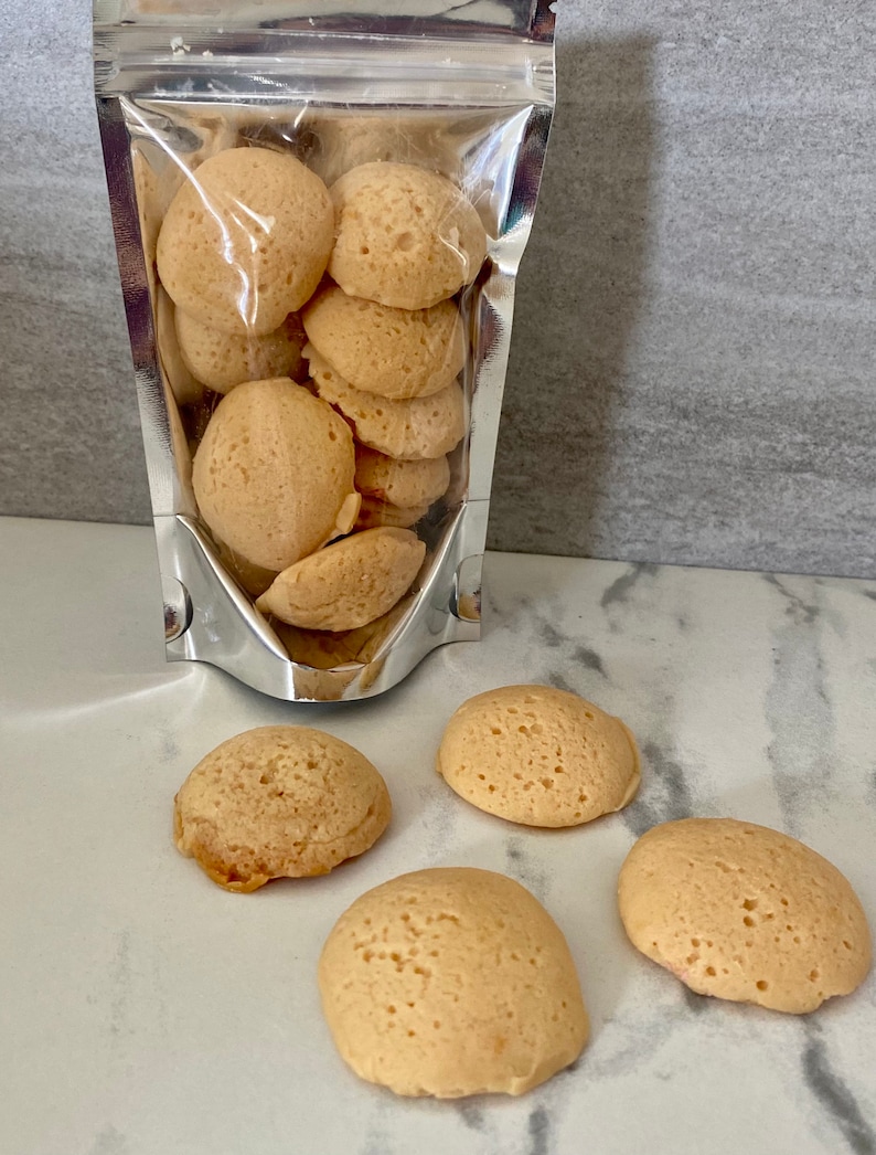 Puede incluir: Una bolsa de cierre herm&eacute;tico plateada llena de galletas redondas y esponjosas de color marr&oacute;n claro. Cuatro galletas adicionales est&aacute;n esparcidas sobre una superficie de m&aacute;rmol blanco.