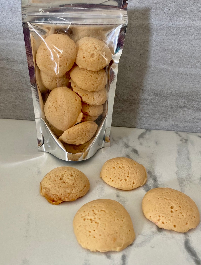 Puede incluir: Una bolsa de cierre herm&eacute;tico plateada llena de galletas redondas y esponjosas de color marr&oacute;n claro. Cuatro galletas adicionales est&aacute;n esparcidas sobre una superficie de m&aacute;rmol blanco.