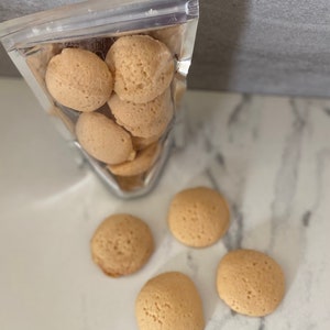 Puede incluir: Una bolsa de pl&aacute;stico transparente llena de galletas redondas de color marr&oacute;n claro. La bolsa est&aacute; abierta y las galletas se est&aacute;n derramando sobre una superficie de m&aacute;rmol blanco.