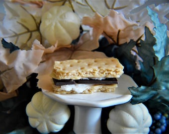 Könnte beinhalten: Ein S'more auf einem weißen Kuchenständer mit weißem Hintergrund. Der S'more besteht aus zwei Graham-Crackern, einer Schokoladen- und einer Marshmallowschicht.