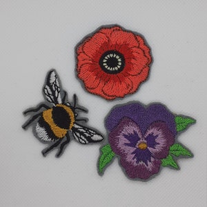 May include: Three embroidered iron-on patches: a red poppy flower, a purple pansy flower, and a bumblebee.