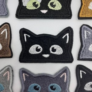 May include: A set of nine embroidered iron-on patches featuring cartoon cats with different coloured fur and eyes. The patches are perfect for adding a touch of whimsy to clothing, bags, or other fabric items.
