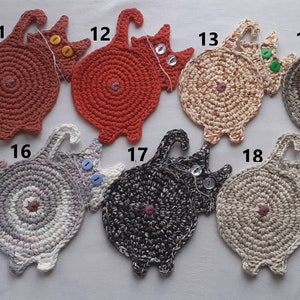 May include: Nineteen crocheted cat-shaped coasters in various colours, including beige, brown, red, grey, and white. Each coaster has a button for an eye and a tail.