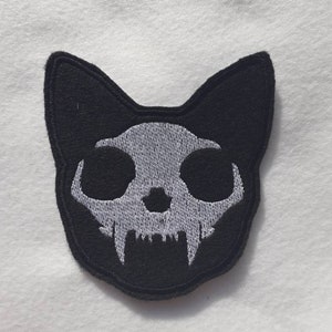 May include: Black embroidered patch of a cat skull with white teeth and eyes.