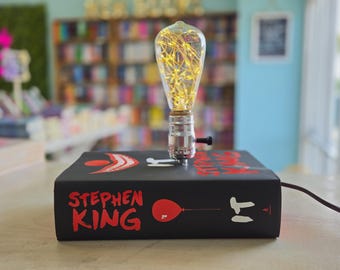 May include: A table lamp constructed from a black hardcover book titled IT by Stephen King, featuring a red balloon graphic and a smiling clown mouth. A silver metal socket holds a clear glass Edison-style light bulb filled with glowing fairy lights.