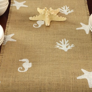 May include: A brown burlap table runner with a white frayed edge. The runner is decorated with white seahorse, starfish, and coral designs.