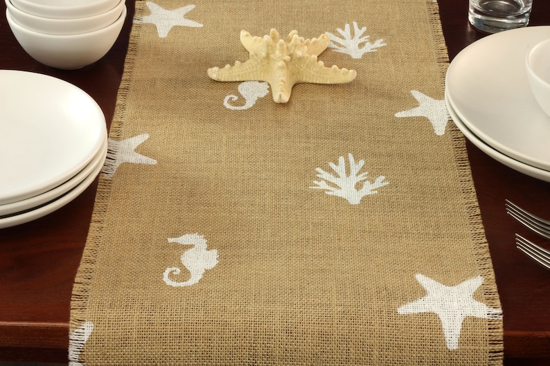 May include: A brown burlap table runner with a white frayed edge. The runner is decorated with white seahorse, starfish, and coral designs.