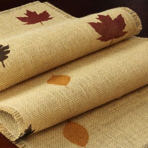 May include: A burlap table runner with a frayed edge features a repeating pattern of brown, orange, and red leaves.