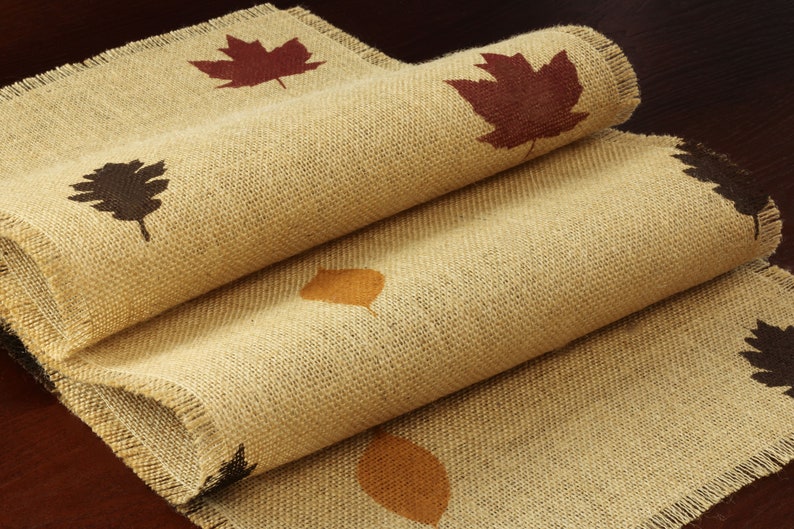 May include: A burlap table runner with a frayed edge features a repeating pattern of brown, orange, and red leaves.