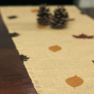 May include: A beige burlap table runner with brown leaf designs. The runner has a natural, rustic look and is perfect for fall decor.