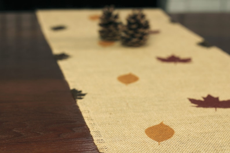 May include: A beige burlap table runner with brown leaf designs. The runner has a natural, rustic look and is perfect for fall decor.