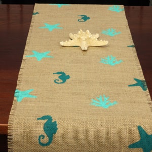 May include: A burlap table runner with a teal blue seahorse, starfish, and coral design. The runner is on a wooden table.