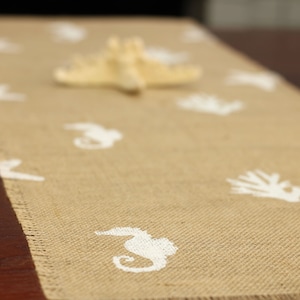 May include: A beige burlap table runner with white seahorse and starfish designs. The runner is on a dark brown wooden table.