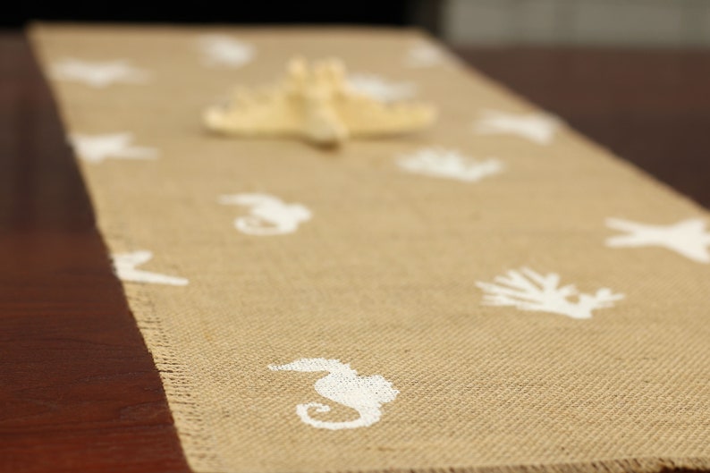 May include: A beige burlap table runner with white seahorse and starfish designs. The runner is on a dark brown wooden table.