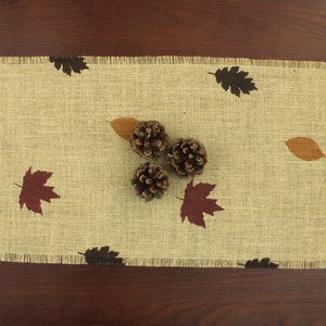 May include: A burlap table runner with a brown, beige, and red leaf pattern. Three pine cones are placed on the runner.