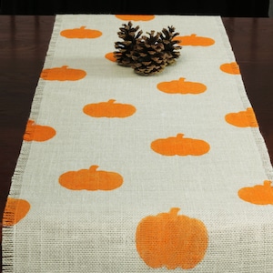 May include: A burlap table runner with a white background and orange pumpkin silhouettes. The runner has frayed edges and is decorated with a pine cone in the center.