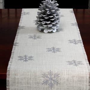 May include: A light beige burlap table runner featuring a repeating pattern of silver snowflakes with frayed edges. A large, silver-painted pinecone is positioned in the center of the runner, resting directly on the textured fabric surface.