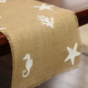 May include: A burlap table runner with a frayed edge features a white starfish, seahorse, and seaweed design.