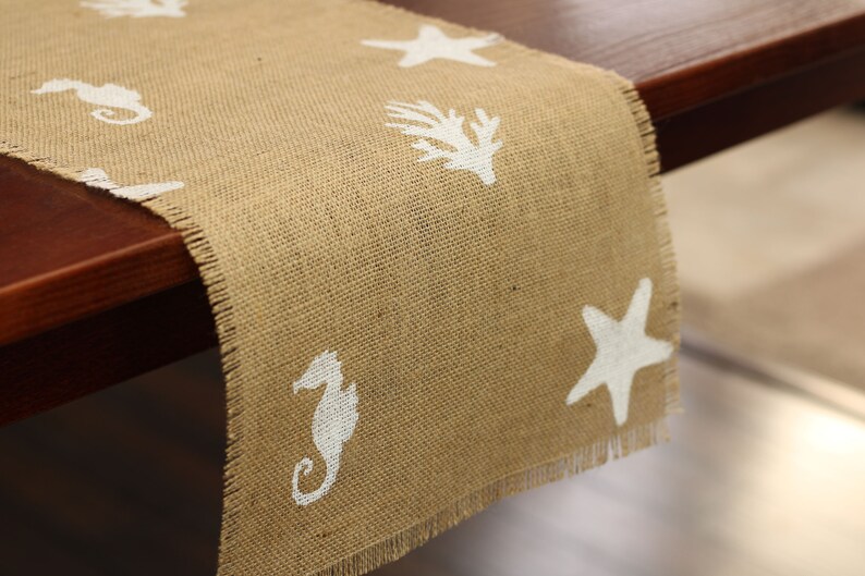 May include: A burlap table runner with a frayed edge features a white starfish, seahorse, and seaweed design.