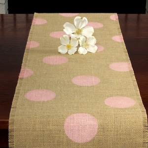 May include: A burlap table runner with pink polka dots. The runner is on a dark wood table with white flowers on top.