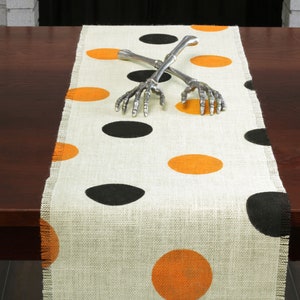May include: A white burlap table runner with black and orange polka dots. Two silver skeleton hands are crossed on top of the runner.