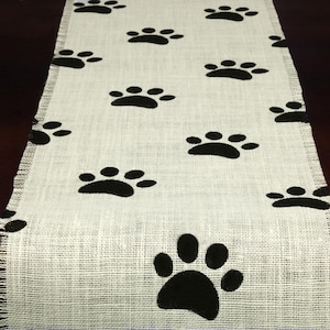 May include: A white burlap table runner with black paw prints. The runner has a frayed edge and is perfect for a dog-themed party.