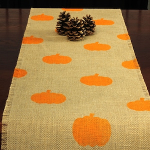 May include: A burlap table runner with orange pumpkin silhouettes. The runner is on a dark wood table with pine cones in the center.