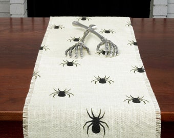May include: A white burlap table runner with a black spider design. Two silver skeleton hand-shaped serving utensils are placed on the runner.
