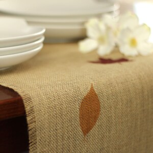 May include: A brown burlap table runner with a frayed edge and a leaf design. The runner is on a wooden table with white plates stacked on top of each other.