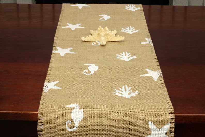 May include: A beige burlap table runner with a white seahorse, starfish, and coral design.