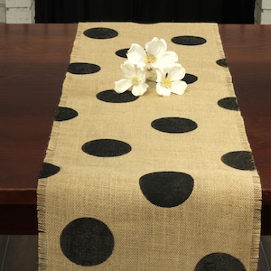 May include: A burlap table runner with black polka dots. The runner is on a dark wood table with white flowers on top.