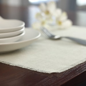 May include: A white placemat with a frayed edge sits on a dark wood table. A stack of white plates and a silver fork are on the placemat.