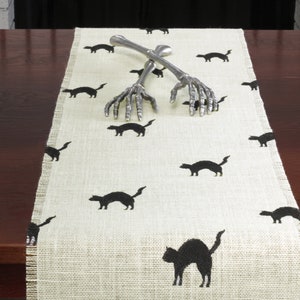 May include: A beige burlap table runner with a black cat design. The runner is decorated with two silver skeleton hand serving utensils.
