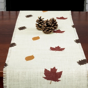 May include: A white burlap table runner with brown and red leaf designs. Two pine cones are placed in the center of the runner.