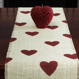 May include: A beige burlap table runner with a red heart pattern. A red heart-shaped candle sits in the center of the runner.