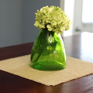 May include: A green glass vase with a wide mouth and a rounded base holds a bouquet of light green hydrangeas. The vase is sitting on a brown burlap placemat on a dark wood table.