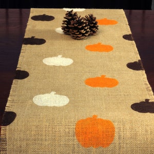 May include: A burlap table runner with a frayed edge features a repeating pattern of orange, white, and brown pumpkins. The runner is decorated with a pine cone in the center.