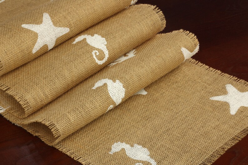 May include: A brown burlap table runner with a frayed edge features a white starfish and seahorse design.